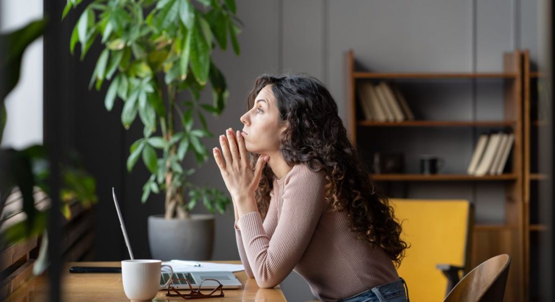 A worker staring pensively into space while seated at her desk in front of her laptop. She is in a modern living room home office.