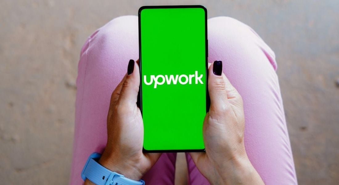 A woman's hands visible in her lap holding a phone with the Upwork logo on its screen on a bright green background. The woman wears pink jeans and a blue watch.