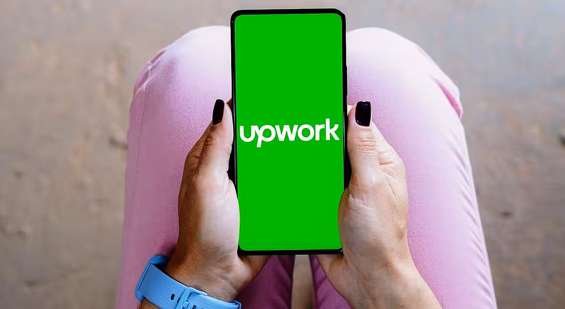 A woman's hands visible in her lap holding a phone with the Upwork logo on its screen on a bright green background. The woman wears pink jeans and a blue watch.