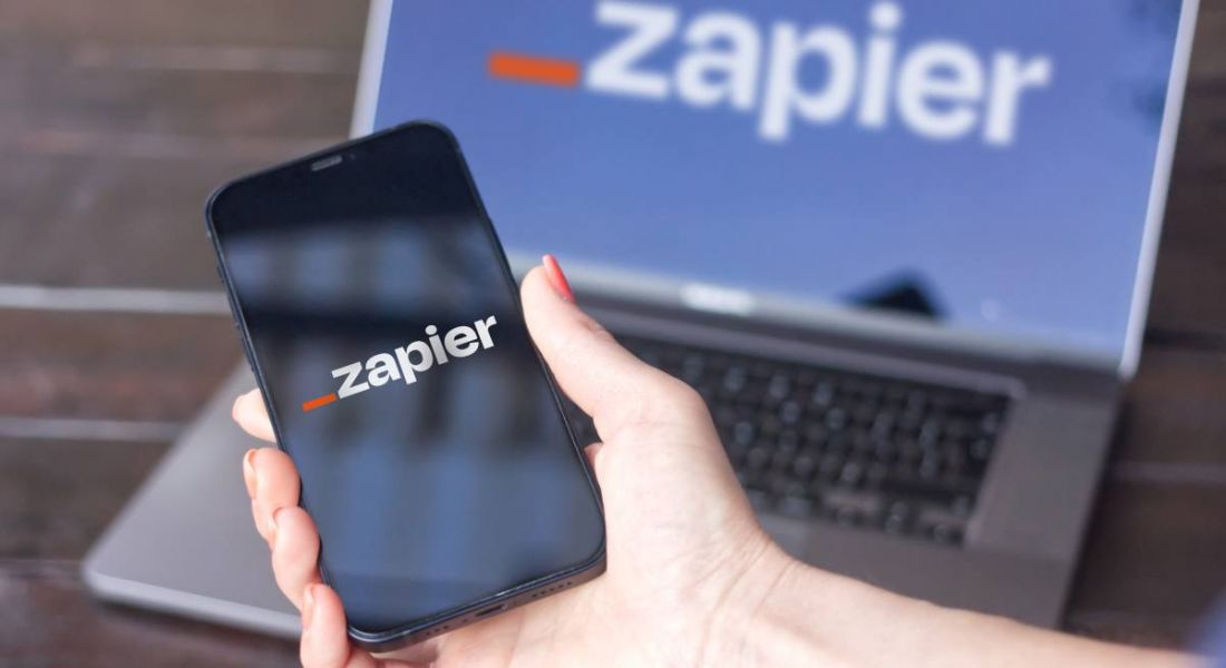 A woman's hand holding a phone with the Zapier logo on the screen. Behind her hand there is a laptop displaying Zapier's logo.