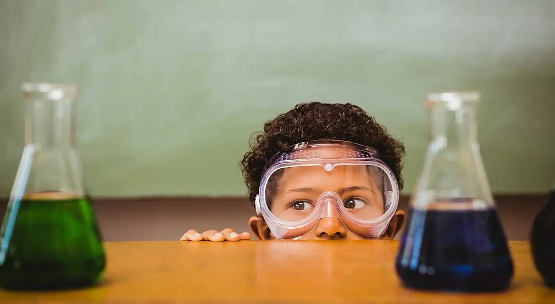 A child wearing protective goggles looks over a tall desk at two beakers containing a green liquid and a blue liquid.