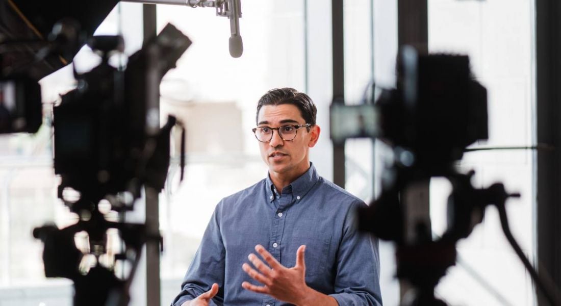 A man wearing glasses and a blue shirt sits in front of two video cameras talking. He is Alex Mahadevan of MediaWise.