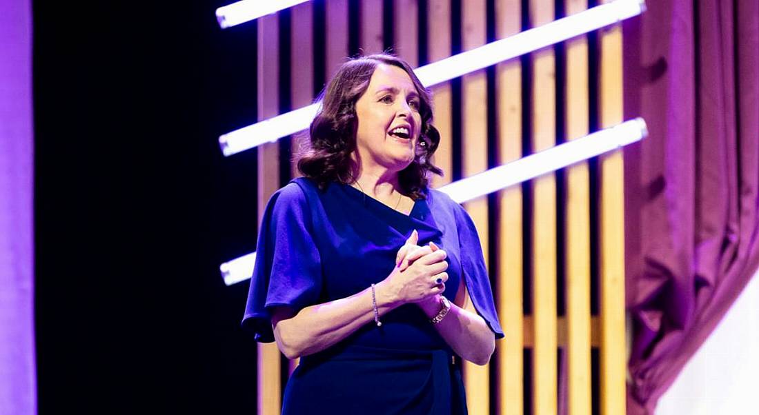 Dr Michelle Cullen of Accenture stands on a stage speaking. There are purple curtains and stage decorations behind her.