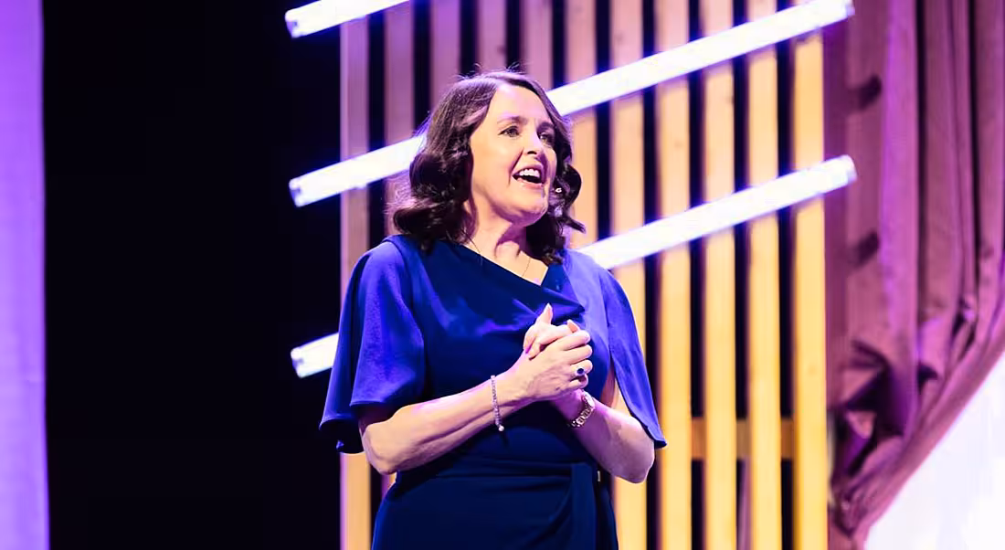 Dr Michelle Cullen of Accenture stands on a stage speaking. There are purple curtains and stage decorations behind her.