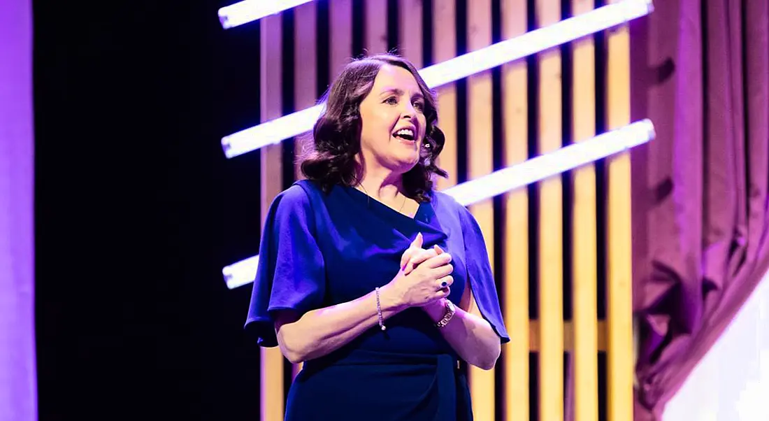 Dr Michelle Cullen of Accenture stands on a stage speaking. There are purple curtains and stage decorations behind her.