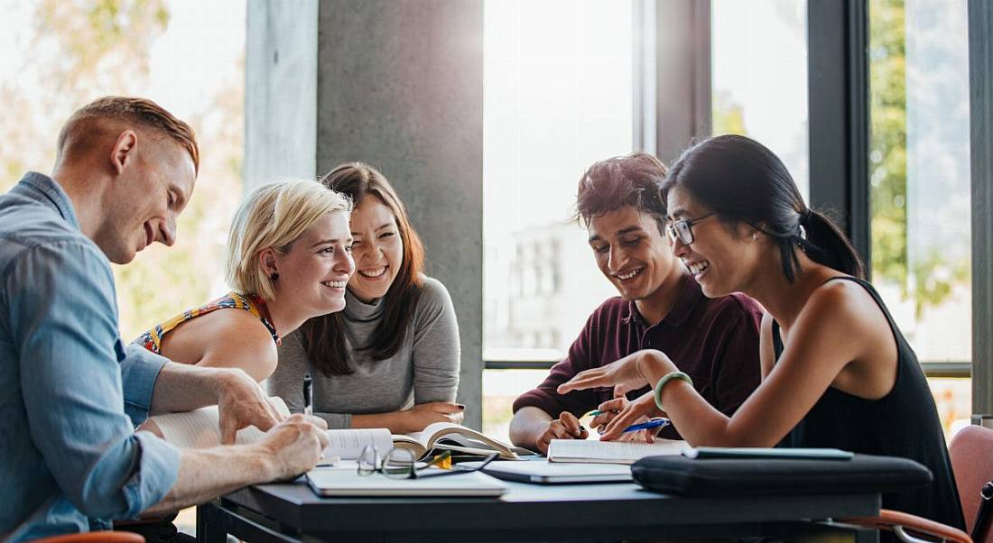 A group of five university students sit around a desk studying and chatting with the sun behind them shining through a window.