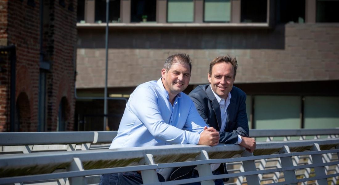 Two men wearing smart casual attire stand side by side leaning on the railing of a bridge on a sunny day.