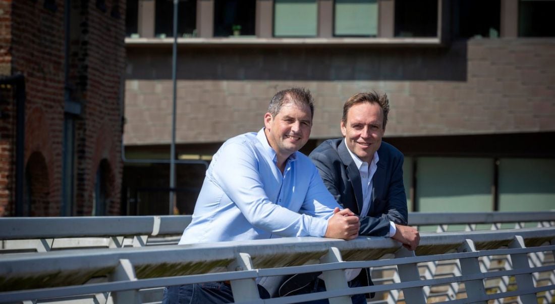 Two men wearing smart casual attire stand side by side leaning on the railing of a bridge on a sunny day.