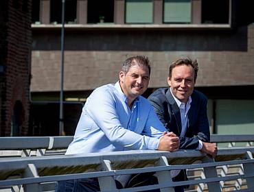 Two men wearing smart casual attire stand side by side leaning on the railing of a bridge on a sunny day.