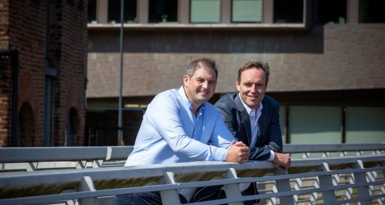 Two men wearing smart casual attire stand side by side leaning on the railing of a bridge on a sunny day.