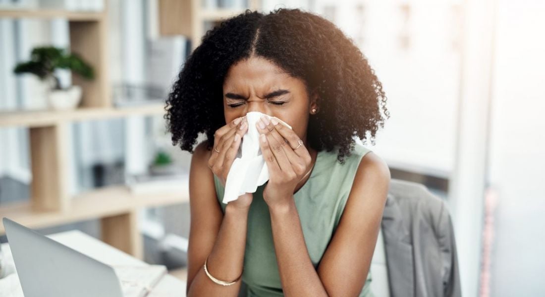 A young black woman at work, blows her nose into a tissue, as she is sick in the office.