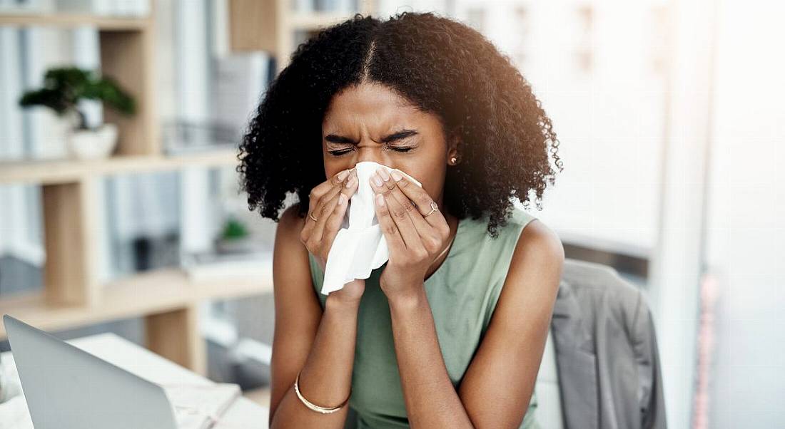 A young black woman at work, blows her nose into a tissue, as she is sick in the office.