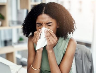 A young black woman at work, blows her nose into a tissue, as she is sick in the office.