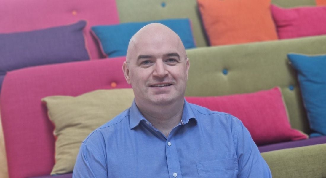 A man wearing a blue shirt smiles while sitting in room surrounded by colourful couches.