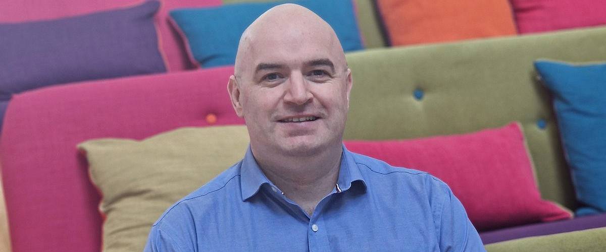 A man wearing a blue shirt smiles while sitting in room surrounded by colourful couches.