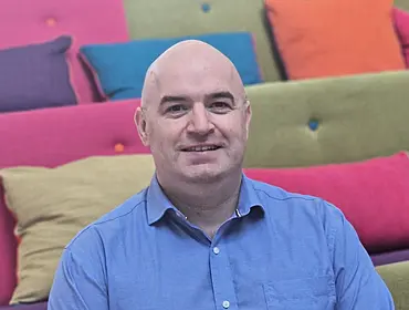 A man wearing a blue shirt smiles while sitting in room surrounded by colourful couches.