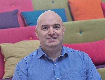 A man wearing a blue shirt smiles while sitting in room surrounded by colourful couches.