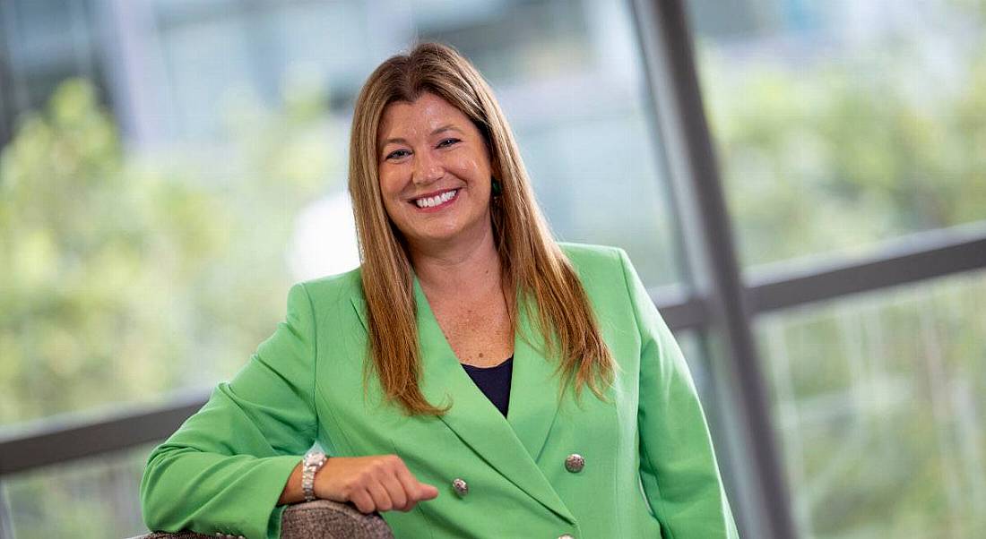 A woman in a green blazer leans on a grey wall in front of an out-of-focus window. She is the MD of Optum UK and Ireland.