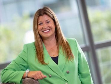 A woman in a green blazer leans on a grey wall in front of an out-of-focus window. She is the MD of Optum UK and Ireland.