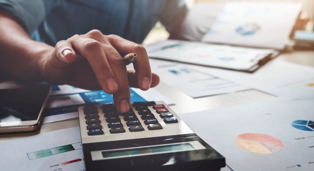 A hand holds a pen and uses a calculator, surrounded by papers as the person works on the budget.