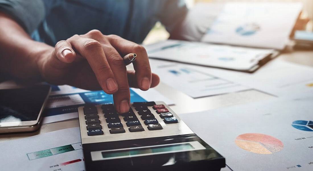 A hand holds a pen and uses a calculator, surrounded by papers as the person works on the budget.