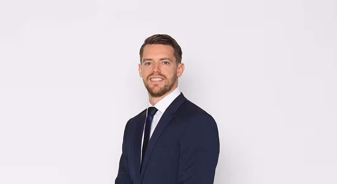 A man in a navy suit smiles at the camera against a plain grey background.