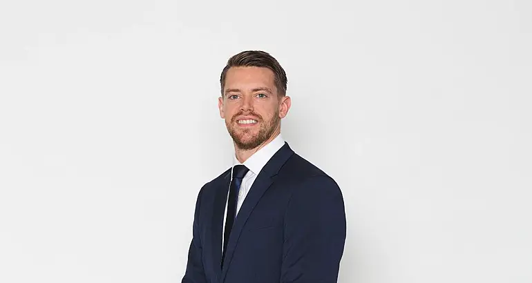 A man in a navy suit smiles at the camera against a plain grey background.