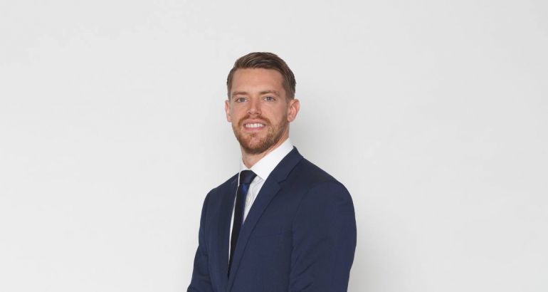 A man in a navy suit smiles at the camera against a plain grey background.