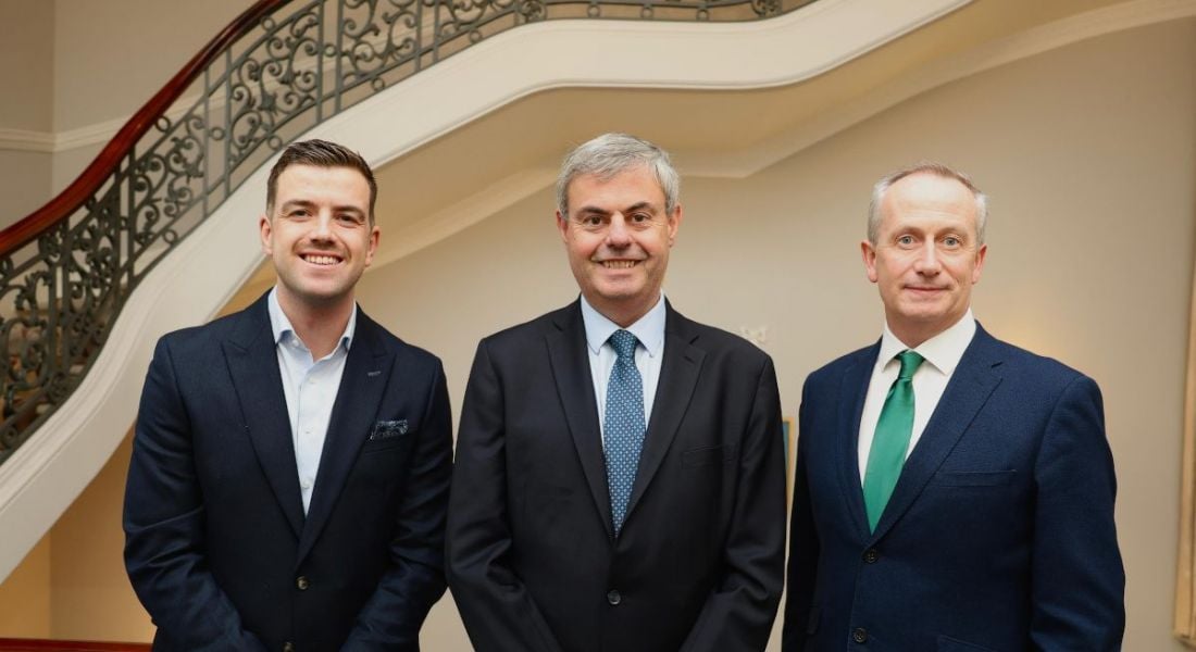 Three men in suits stand side by side smiling at the camera in front of a staircase.