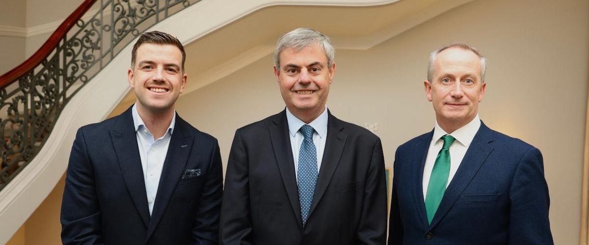 Three men in suits stand side by side smiling at the camera in front of a staircase.