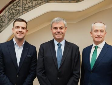 Three men in suits stand side by side smiling at the camera in front of a staircase.