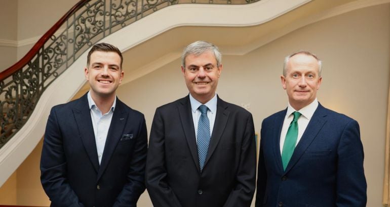 Three men in suits stand side by side smiling at the camera in front of a staircase.