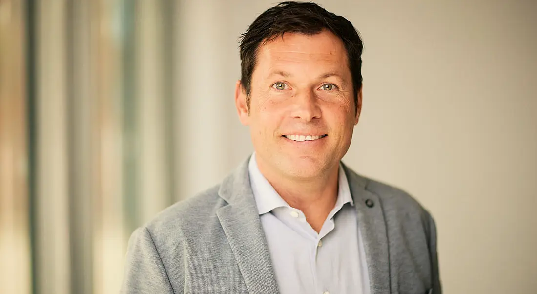 A headshot of Tim Petzinna, a man in a grey suit, smiling at the camera with a soft-focus light background.