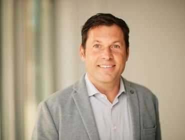 A headshot of Tim Petzinna, a man in a grey suit, smiling at the camera with a soft-focus light background.