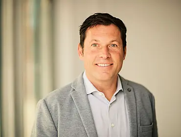 A headshot of Tim Petzinna, a man in a grey suit, smiling at the camera with a soft-focus light background.