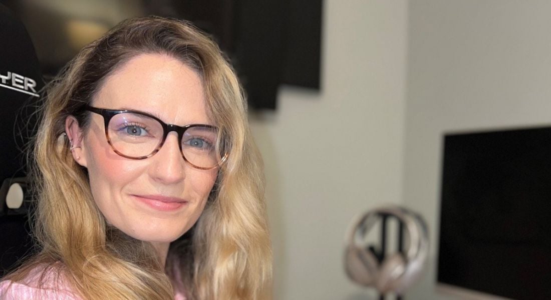 A woman with long blonde hair smiles at the camera while wearing glasses. In the background is a home office set up, with a computer screen seen to the left of the image.