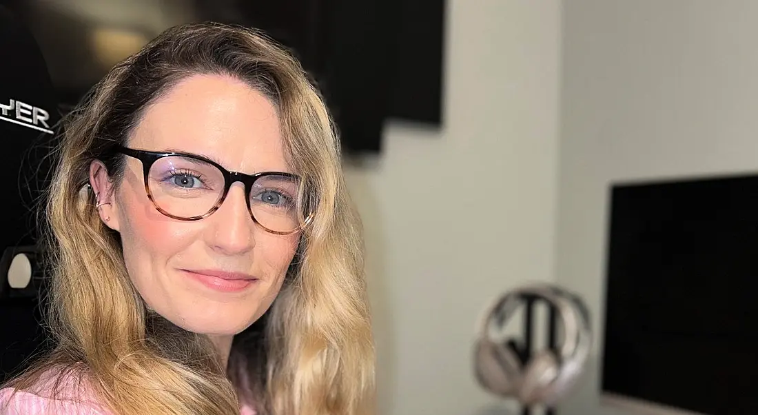 A woman with long blonde hair smiles at the camera while wearing glasses. In the background is a home office set up, with a computer screen seen to the left of the image.