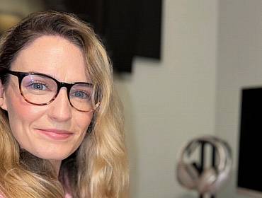 A woman with long blonde hair smiles at the camera while wearing glasses. In the background is a home office set up, with a computer screen seen to the left of the image.