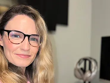 A woman with long blonde hair smiles at the camera while wearing glasses. In the background is a home office set up, with a computer screen seen to the left of the image.