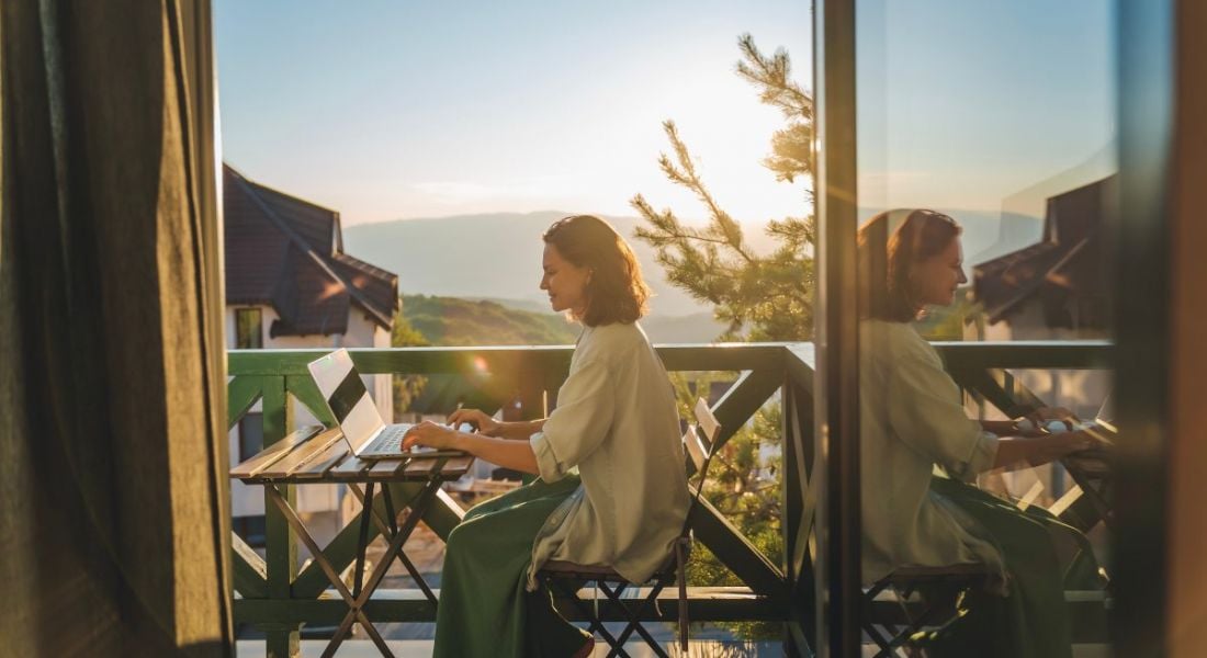 A young woman sits working on a laptop remotely working, with a sunset and mountains on the background.