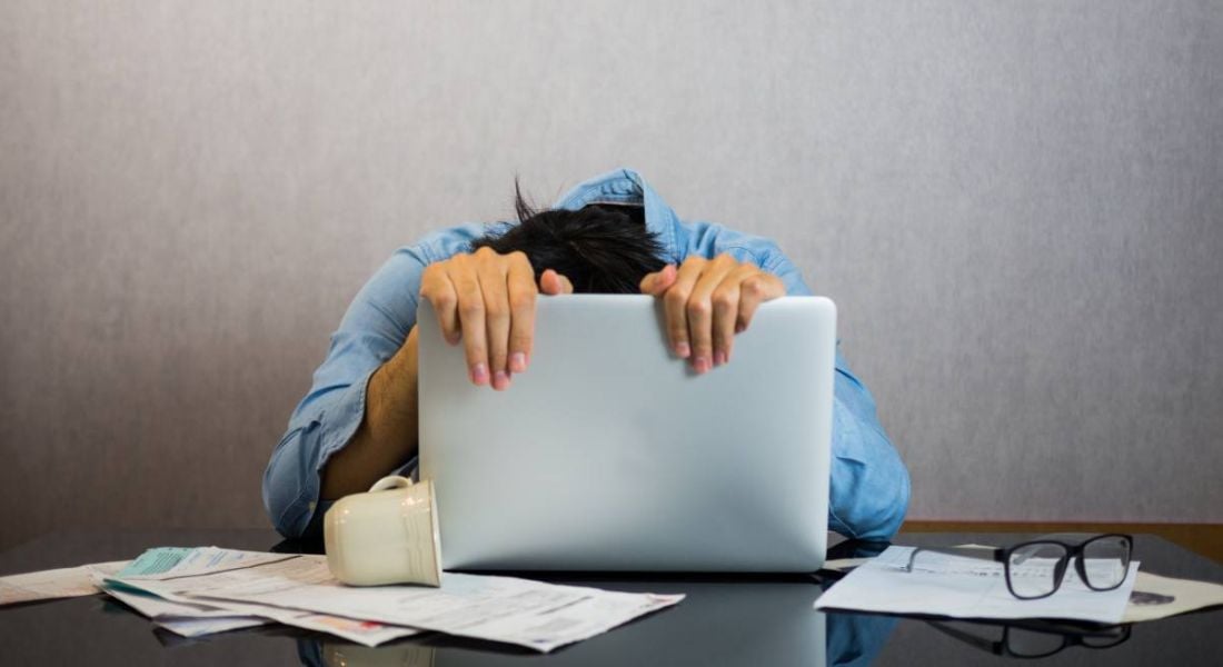 A man clutching his laptop screen with his head down at his desk.