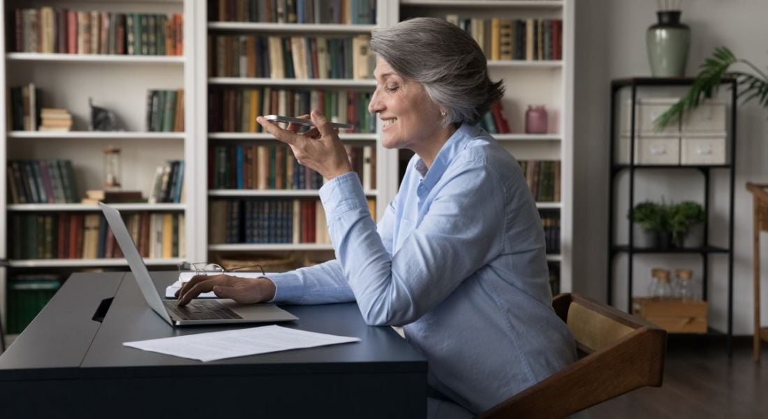 An older employee uses her phone to assist her in her work while sitting at her desk.