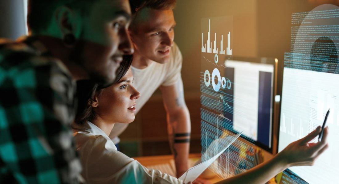 Three people look at two large computer screens. They are software developers and there are data visualisation overlaid on the image.