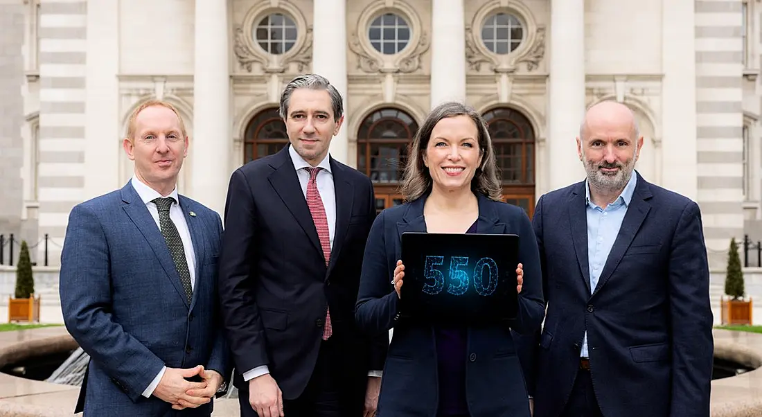 Three men and a woman in business attire stand in a line. The woman holds a tablet that shows the number 550 in blue writing.