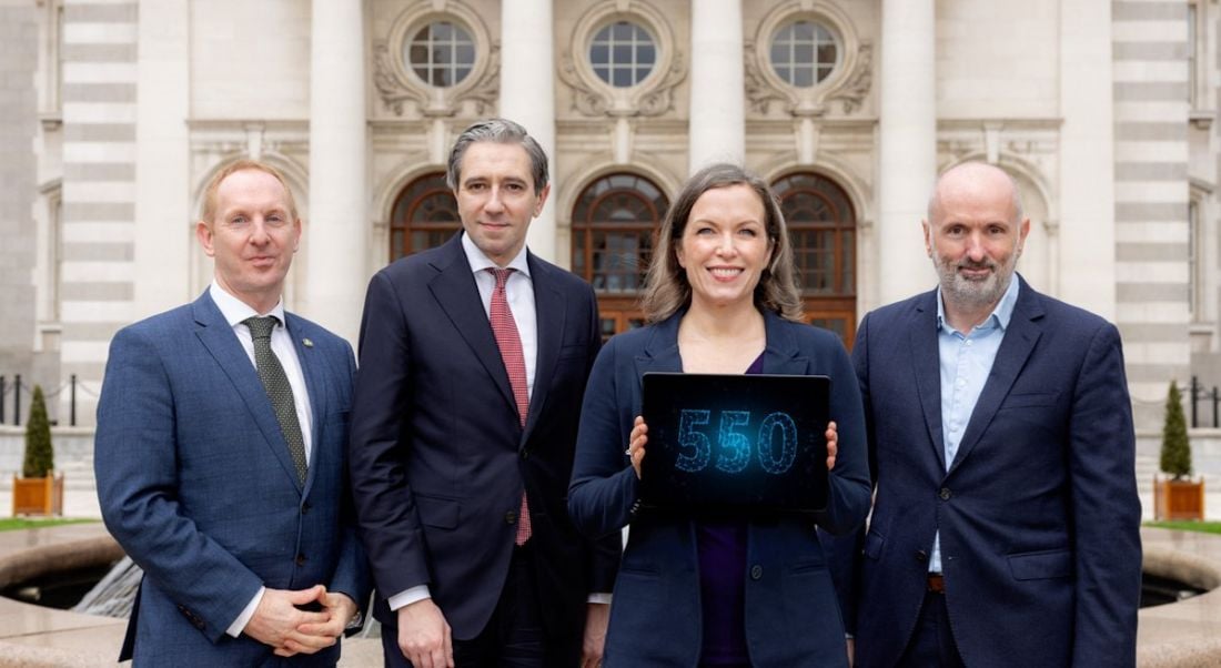 Three men and a woman in business attire stand in a line. The woman holds a tablet that shows the number 550 in blue writing.