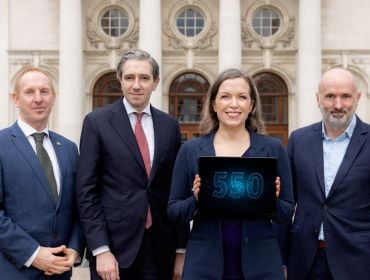 Three men and a woman in business attire stand in a line. The woman holds a tablet that shows the number 550 in blue writing.