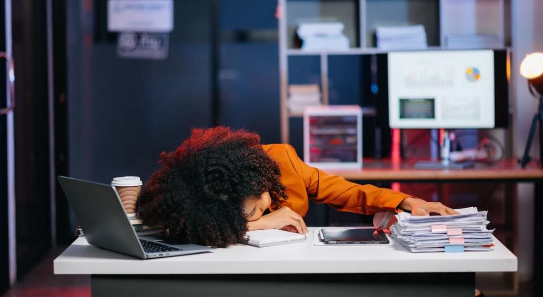 A young black woman dressed in orange is slumped at her desk, exhausted.