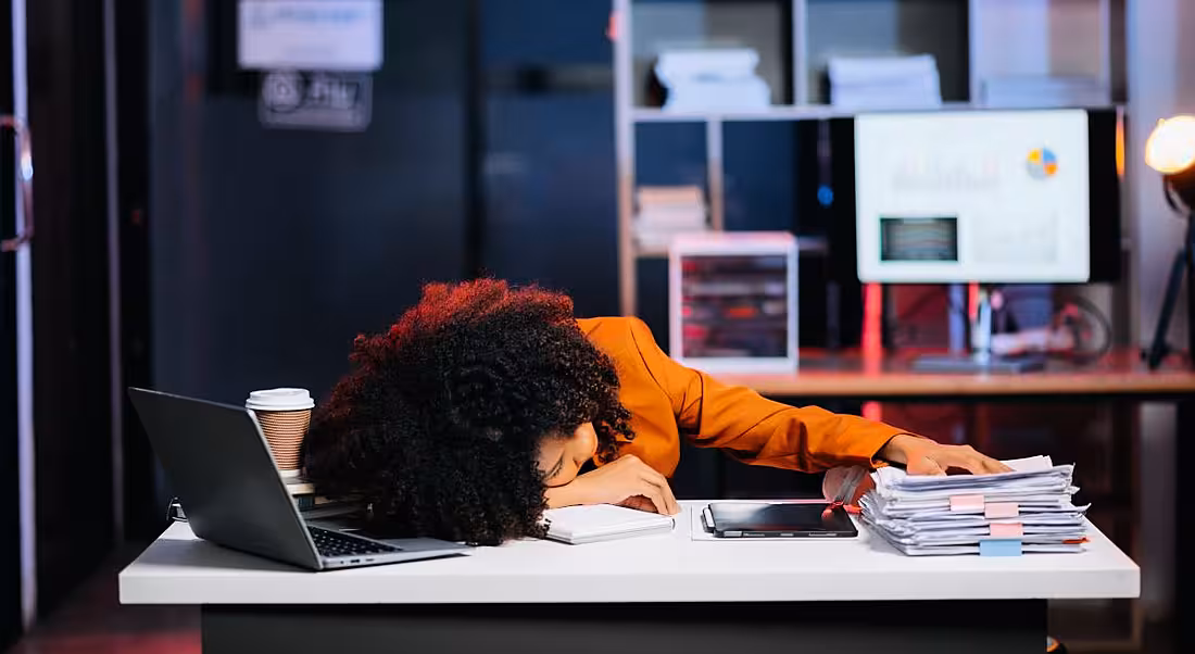 A young black woman dressed in orange is slumped at her desk, exhausted.