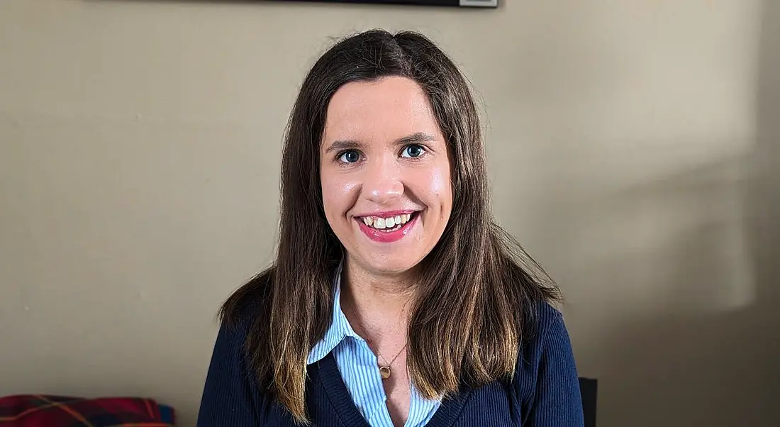 A woman with long brown hair smiles at the camera while sitting in front of a beige wall.