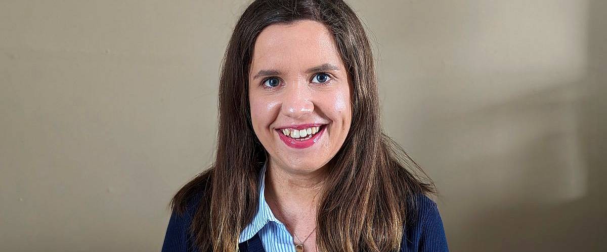 A woman with long brown hair smiles at the camera while sitting in front of a beige wall.